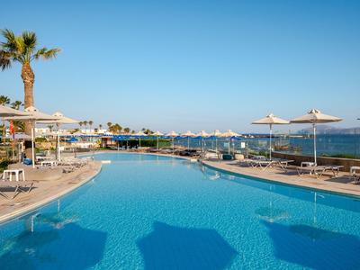 Großer Hotelpool mit Liegestühlen und Sonnenschirmen neben Meer und blauem Himmel.
