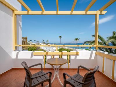 Balkon mit Tisch, zwei Stühlen unter gelber Pergola mit Blick auf tropische Palmen bei klarem Himmel.