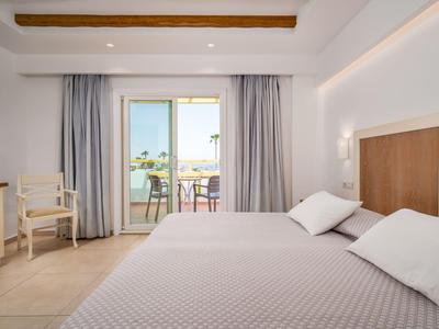 Helles Hotelzimmer mit großem Bett, weißer Bettwäsche und Blick auf Balkon mit Meer.