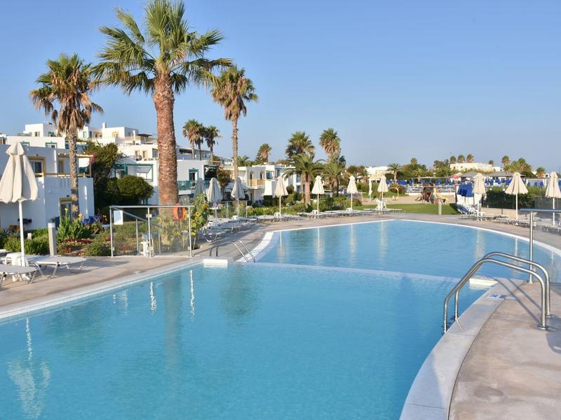 Großer Swimmingpool mit Palmen und weißen Sonnenschirmen in einer Hotelanlage bei klarem Himmel.