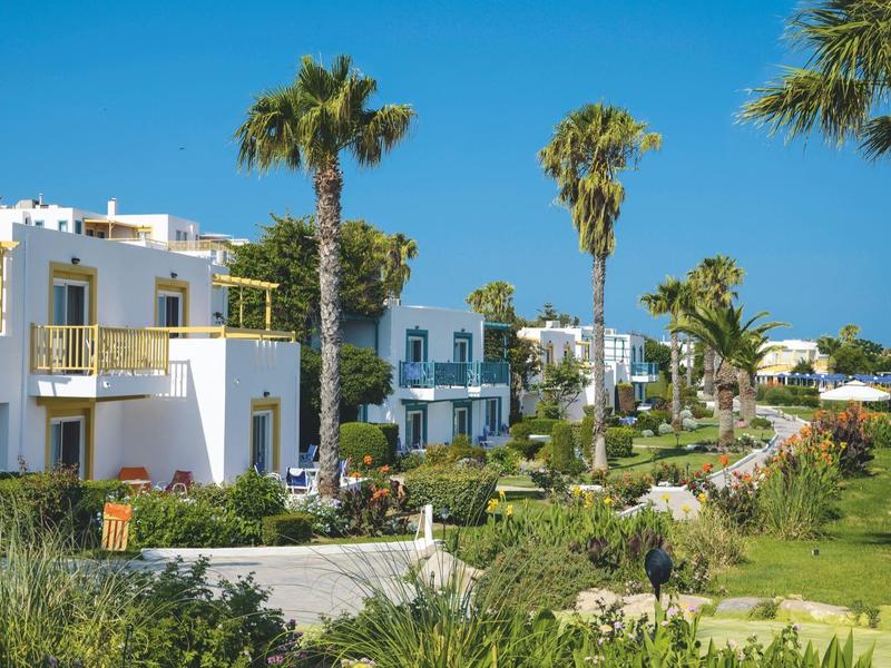 Reihen von weißen Häusern mit Balkonen und Palmen in einer gepflegten Gartenanlage bei blauem Himmel.