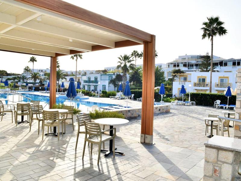 Sonniges Poolareal mit weißen Tischen und Stühlen, Palmen und weißen Gebäuden im Hintergrund.