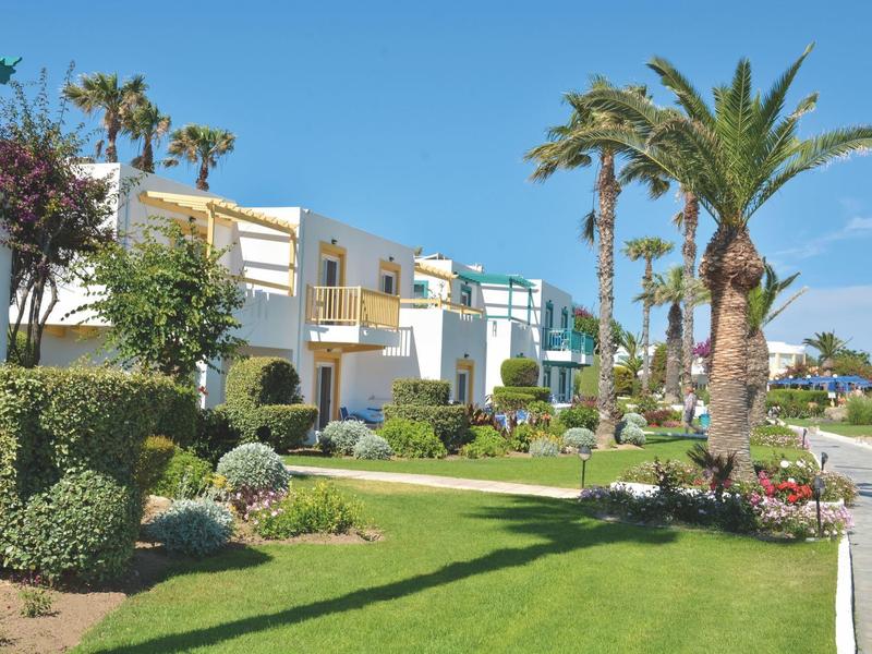 Mehrere moderne weiße Reihenhäuser mit grünen Gärten und Palmen bei klarem Himmel.