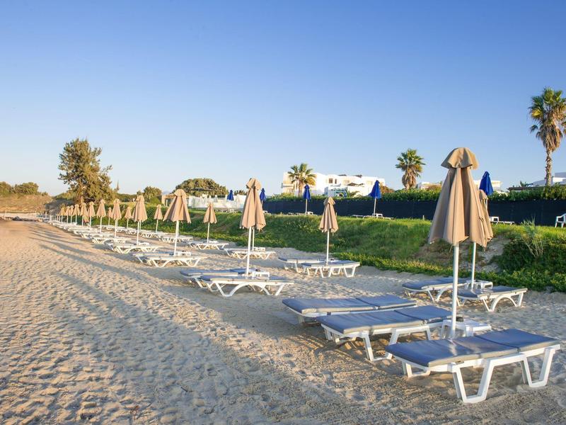Leere Sonnenliegen mit geschlossenen Sonnenschirmen am Strand bei bewölktem Himmel.
