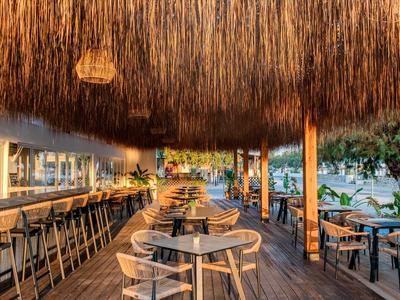 Otwarta przestrzeń restauracyjna z drewnianą podłogą, strzechą i nowoczesnymi stolikami oraz krzesłami.