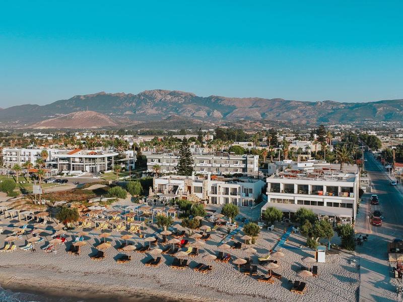Strand mit Sonnenschirmen und Hotels davor, im Hintergrund Berge unter klarem blauem Himmel.