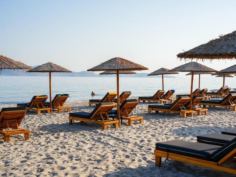 Strand mit Holzliegen und Sonnenschirmen aus Stroh vor ruhigem, blauem Meer und klarem Himmel.