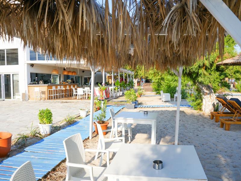 Strandrestaurant mit Tischen, weißen Stühlen und Strohdach, Sandboden und Sonne im Hintergrund.