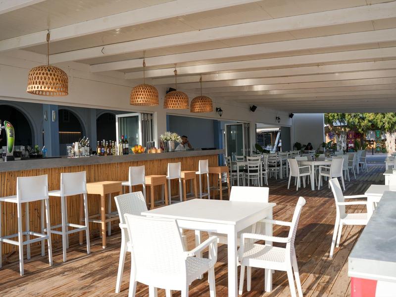 Moderner Außenbereich mit Bar, weißen Stühlen und Tischen, Holzboden und geflochtenen Lampen.