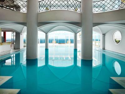 Vnitřní bazén s bílými sloupy a velkými obloukovými okny odrážejícími se v čisté modré vodě.