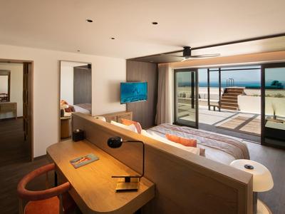 Moderní hotelový pokoj s psacím stolem, postelí, televizí a skleněnými dveřmi na balkon s výhledem na pláž.