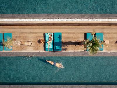 Letecký pohled na ženu plavící se v bazénu u tří lehátek s palmami.