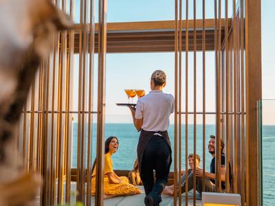 Číšník podává nápoje dvěma osobám sedícím na dřevěné terase u moře.