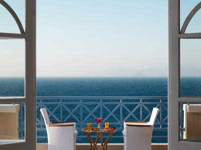 Dva židle a nápoje na stole čelí moři z balkonu s zábradlím.