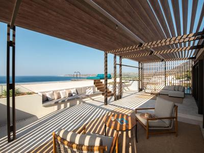 Zastíněná terasa s posezením s výhledem na písečnou pláž a klidné moře pod jasnou oblohou.