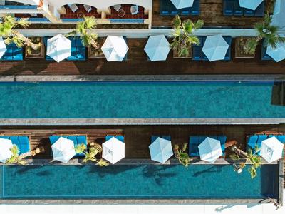 Pohled shora na hotelový bazén se dvěma prodlouženými obdélníkovými bazény oddělenými úzkou chodníkem s deštníky a lehátky.