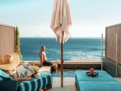 Relaxter Poolbereich mit Blick auf das Meer, zwei entspannte Personen und eine geschlossene Sonnenschirm.