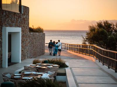 Drei Personen gehen zum Meer bei Sonnenuntergang neben einem Steinhaus und Outdoor-Sitzbereich.