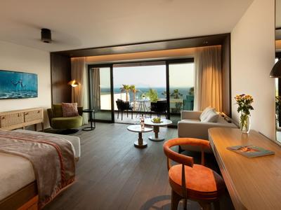 Habitación de hotel moderna con ventana grande, vista al mar, cama doble y zona de estar acogedora.