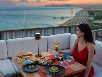 Mujer con vestido rojo sentada en una mesa con comida en una terraza con vista al mar al atardecer.