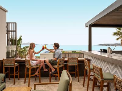 Pareja sentada en barra con vista al mar, brindando en terraza soleada del hotel.