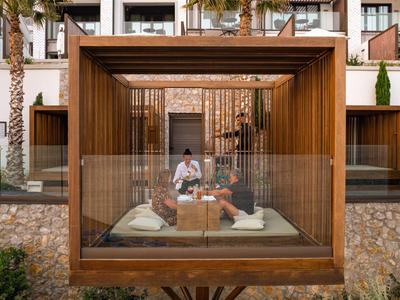 Balcón moderno de hotel con zona de estar de madera y grandes paneles de cristal, rodeado de palmeras.