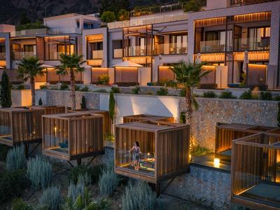 Hotel moderno en una ladera con terrazas de madera iluminadas y montañas al fondo al atardecer.