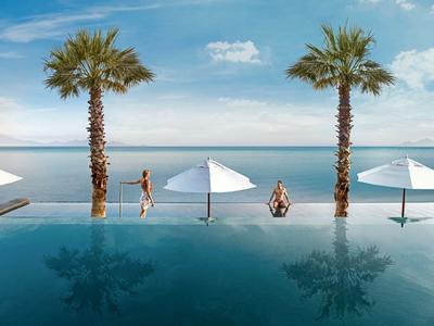 Piscina infinita con palmeras y sombrillas blancas junto al mar en un día soleado.