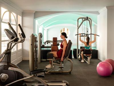 Dos personas entrenan en un gimnasio luminoso con equipo cardiovascular y de fuerza.