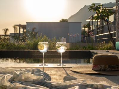 Dos vasos sobre el borde de una piscina al atardecer, junto a un sombrero y una tela.