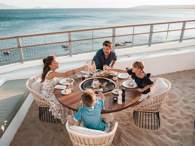 Familia sentada en una mesa redonda sobre arena con vista al mar.