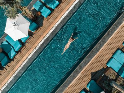 Mujer nadando en una piscina estrecha rodeada de tumbonas azules y una sombrilla blanca.