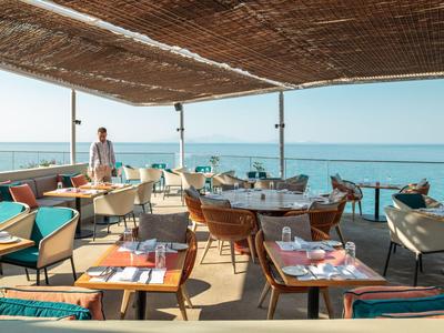 Restaurante abierto junto al mar con techo de madera, mesas de madera y sillas coloridas a la luz del día.