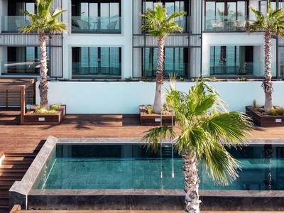 Piscina moderna de hotel con terraza de madera y palmeras frente a un edificio de varios pisos.