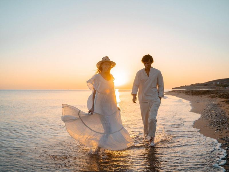 Pár v bílém kráčí po pláži při západu slunce s jemnými oceánskými vlnami.