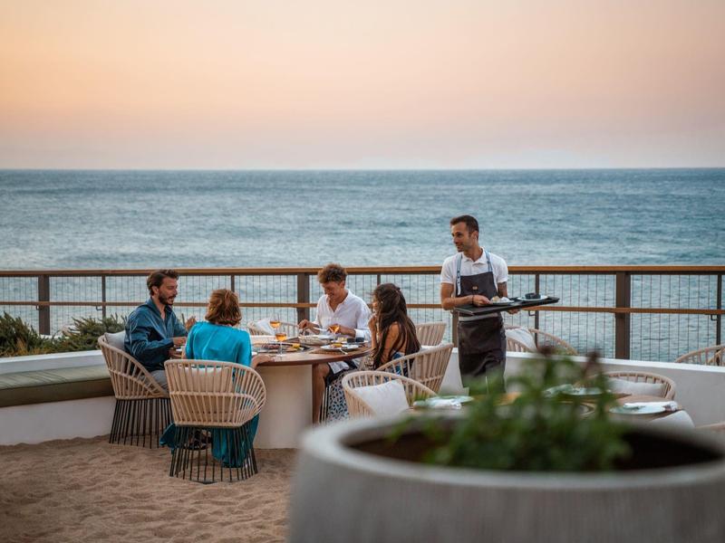 Hosté večeří venku při západu slunce u moře, číšník podává jídlo na terase.