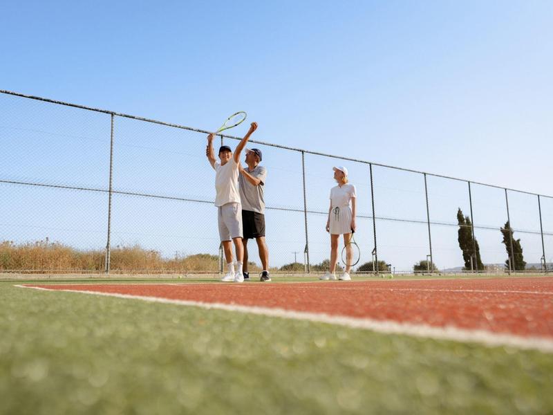 Lidé hrají tenis u oploceného kurtu pod jasnou modrou oblohou.