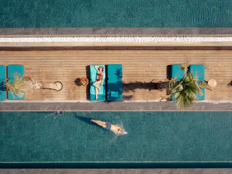 Letecký pohled na ženu plavící se v bazénu u tří lehátek s palmami.