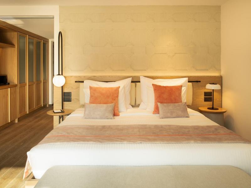 Pohodlný hotelový pokoj s manželskou postelí, polštáři a otevřenou skříní v teplém osvětlení.