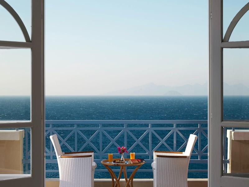 Dva židle a nápoje na stole čelí moři z balkonu s zábradlím.