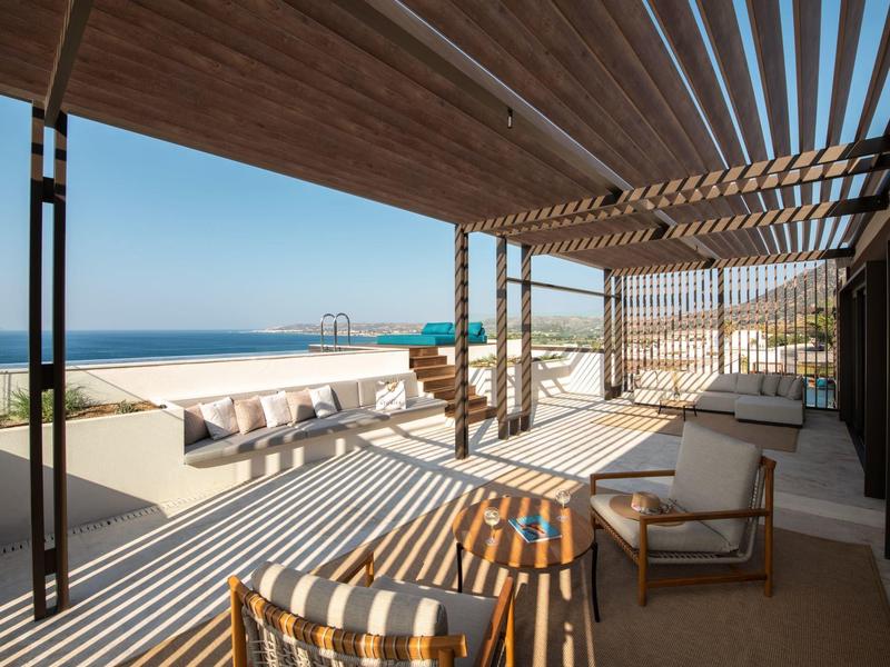 Zastíněná terasa s posezením s výhledem na písečnou pláž a klidné moře pod jasnou oblohou.