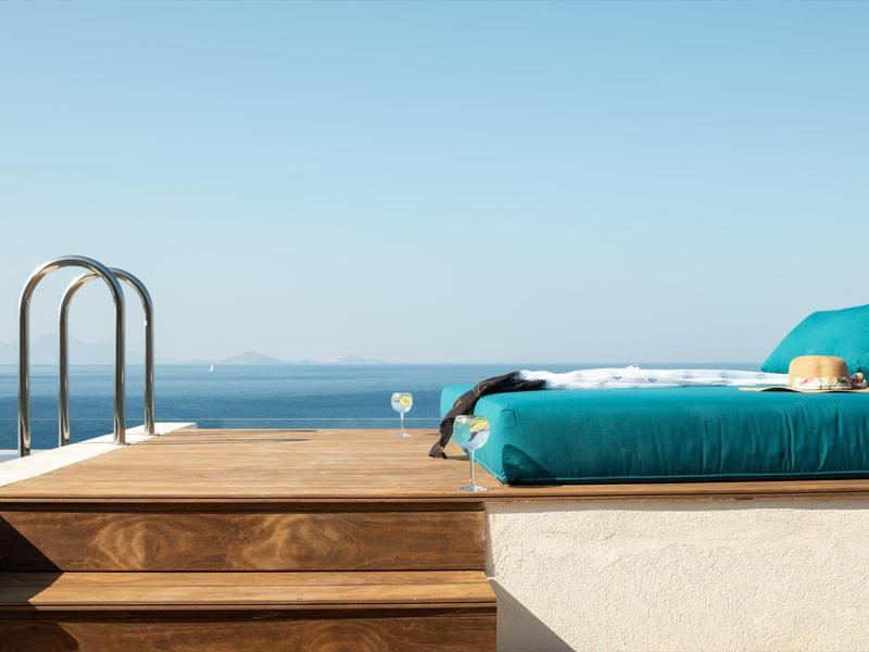 Bazén s schody a lehátky s tyrkysovými polštáři, výhled na moře za jasné oblohy.