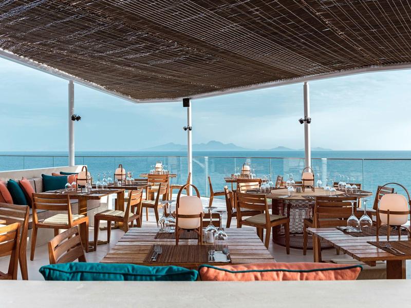 Venkovní terasa restaurace s dřevěnými stolky a výhledem na moře pod bambusovou střechou.