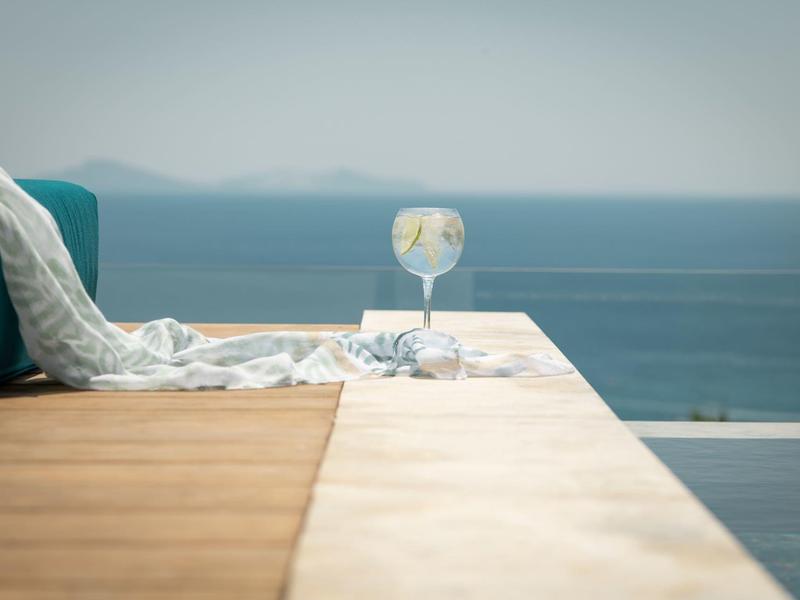 Sklenice na koktejl stojí na okraji bazénu s výhledem na moře a jasnou oblohu.