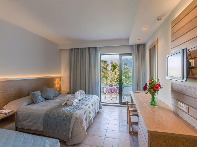 Světle hotelový pokoj se dvěma postelemi, psacím stolem, terasou a výhledem na palmy.