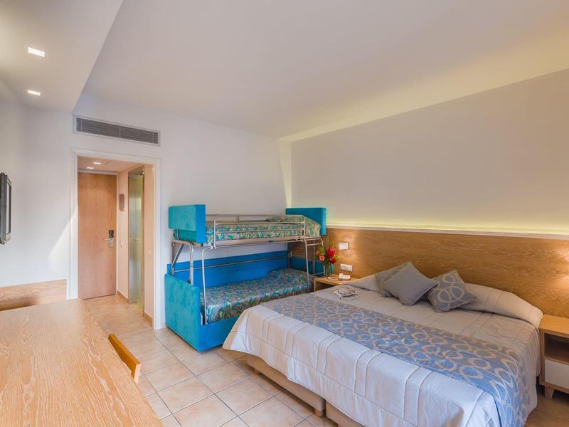 Moderní hotelový pokoj s manželskou postelí, patrovou postelí a světlou dřevěnou podlahou.