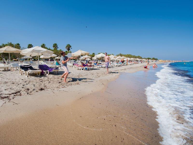 Slunná pláž s slunečníky, lehátky a lidmi vedle klidného modrého moře.