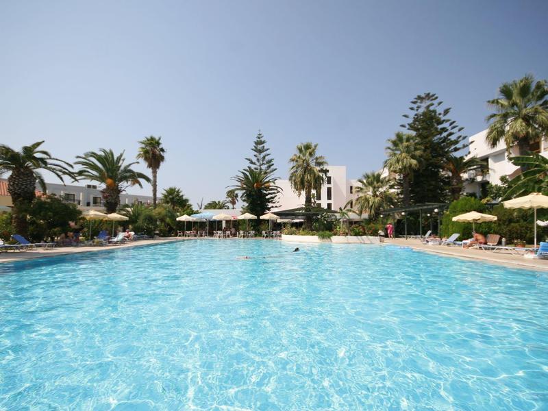 Velký modrý hotelový bazén s lehátky a palmami pod jasnou oblohou.