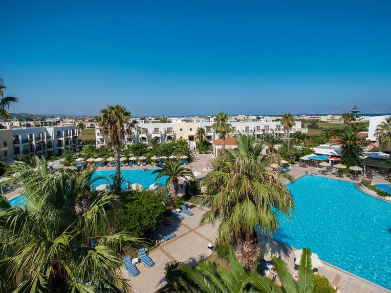 Velké venkovní bazény obklopené palmami a bílými hotelovými budovami pod jasně modrou oblohou.