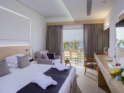 Světelný hotelový pokoj s manželskou postelí, křeslem a balkonem s výhledem na moře.